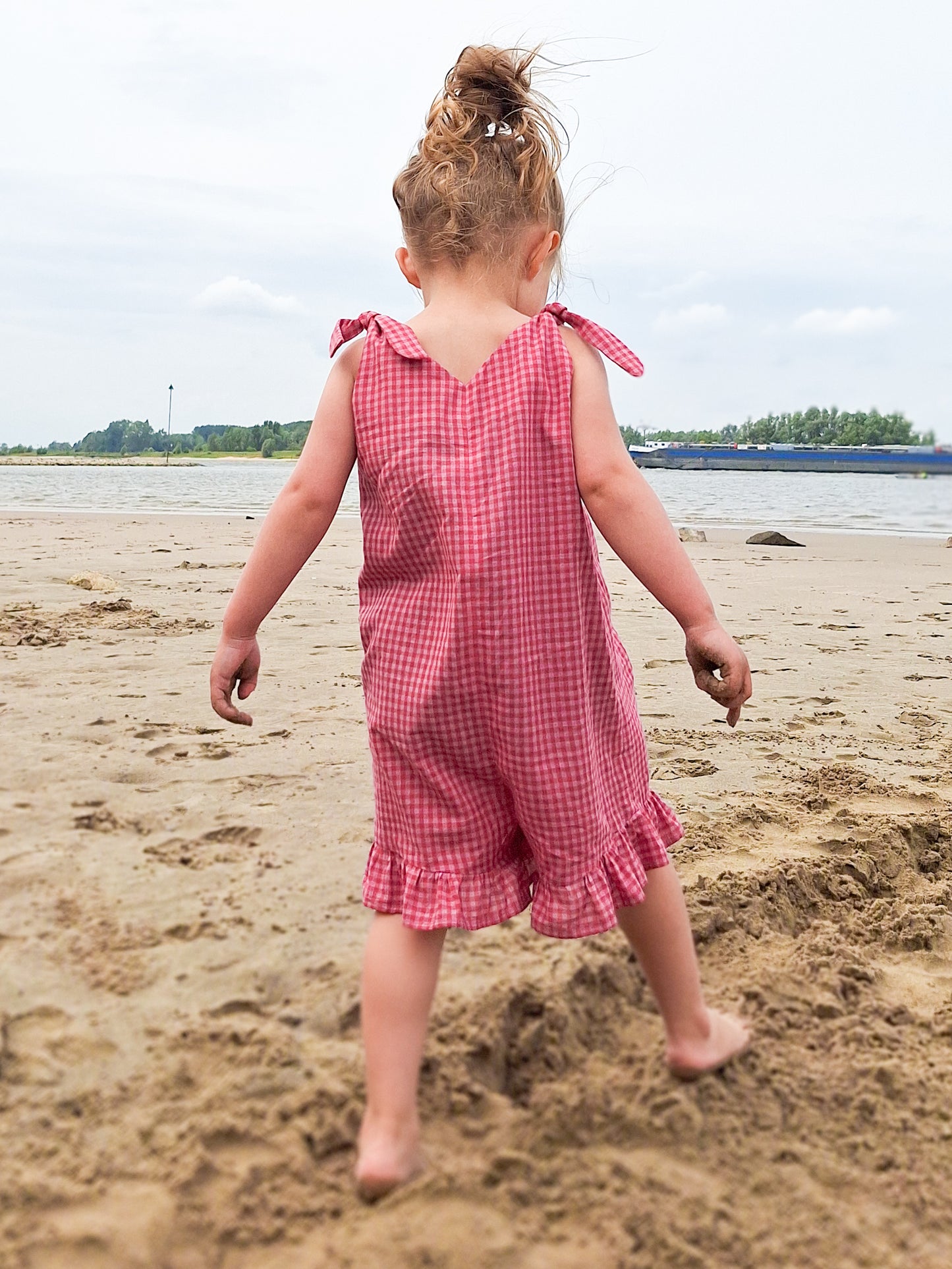 Child in a pink checkered dress standing on a sandy beach.