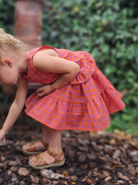 Child wearing a pink checkered dress standing outdoors on mulch with greenery in the background
