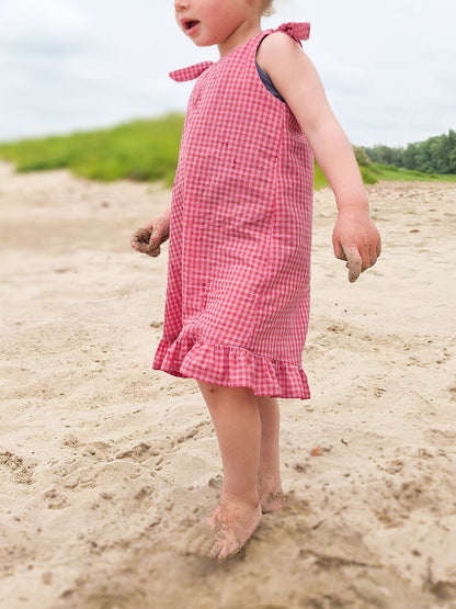 Child in a pink checkered dress standing on a sandy beach.