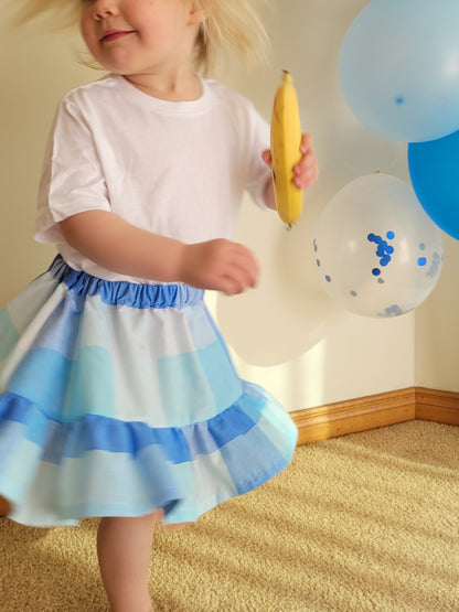 Child in a white shirt and blue striped skirt holding a banana with balloons in the background.