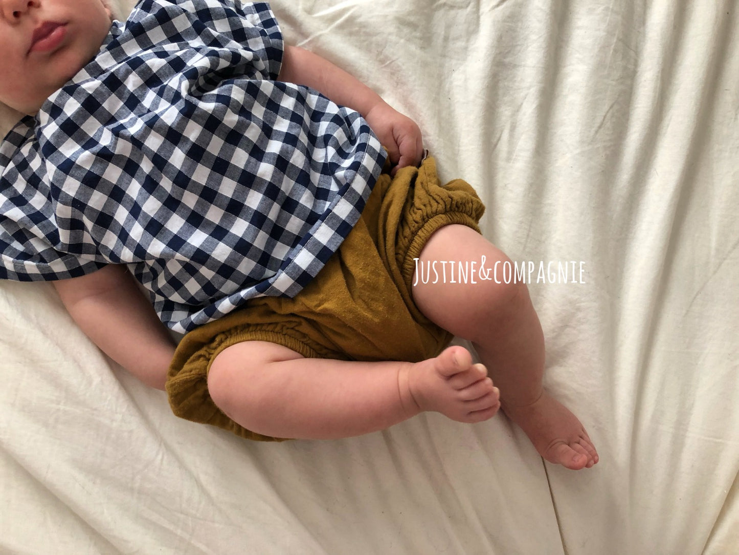 Baby wearing a checkered shirt and brown bloomers on a white blanket, with 'Justine & Compagnie' branding.