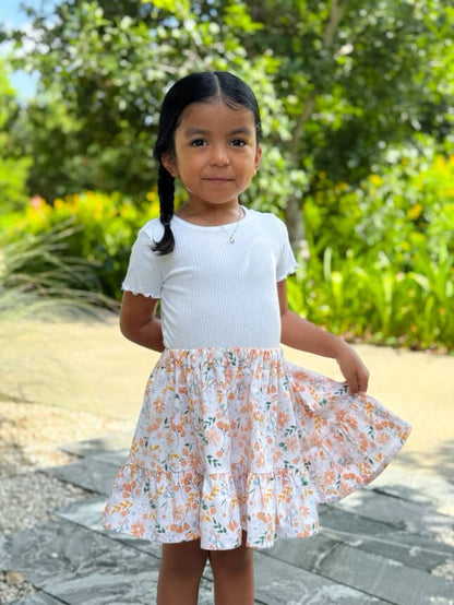 Young girl in a white top and floral skirt standing outdoors with greenery in the background