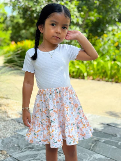 Young girl wearing a white top and floral skirt outdoors with greenery in the background