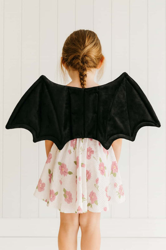 Child wearing black bat wings over a floral dress against a white background