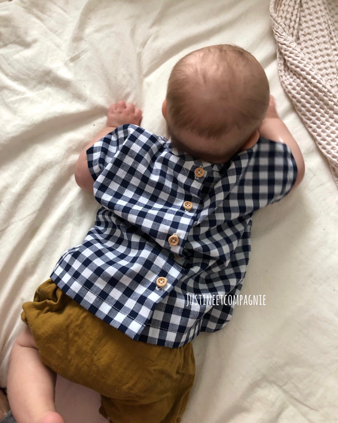Baby wearing a blue and white checkered shirt and mustard yellow shorts on a white blanket.