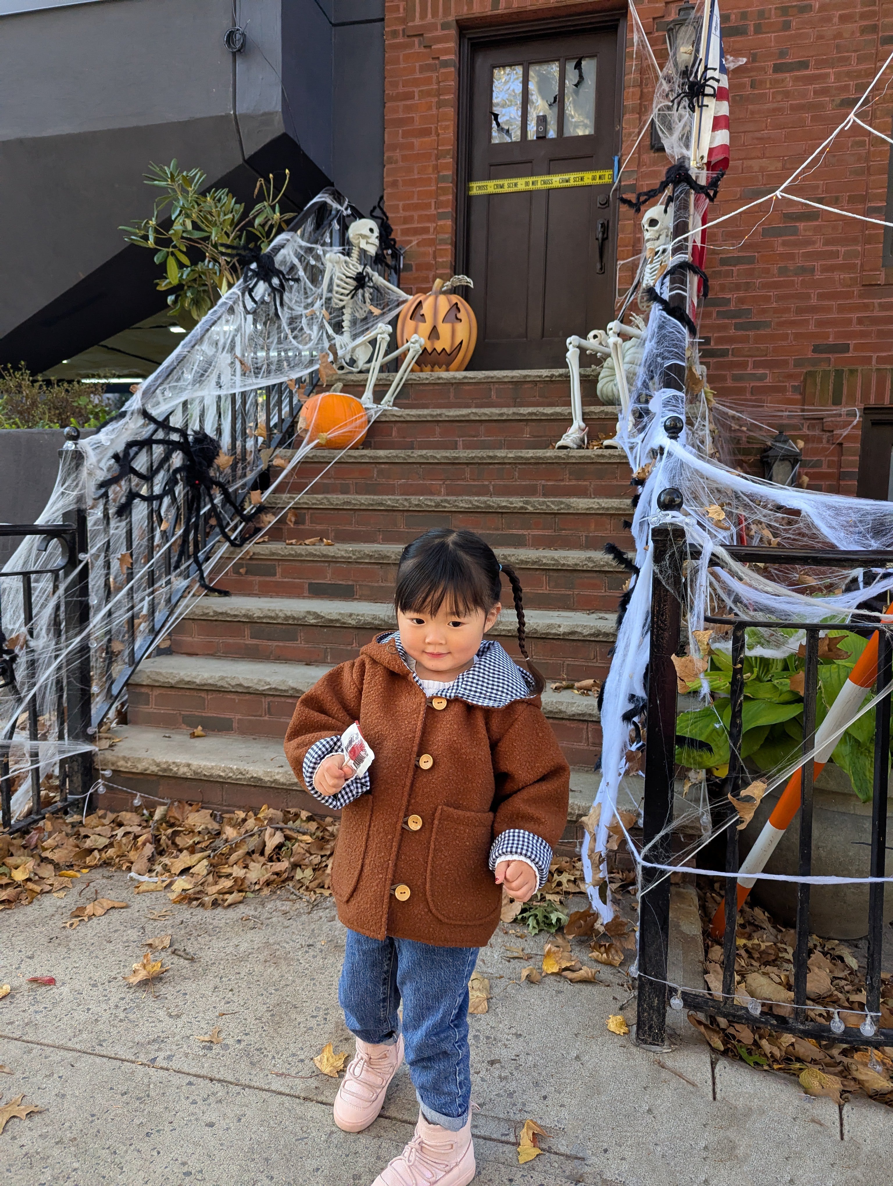 Child in a brown coat standing in front of Halloween-decorated steps with pumpkins and skeletons.