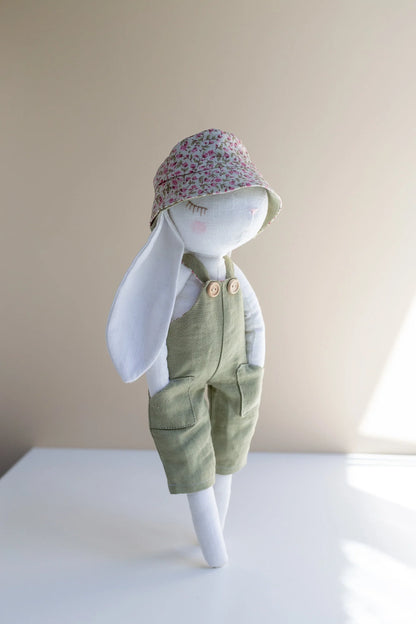 Stuffed bunny toy wearing a floral hat and green overalls on a plain background