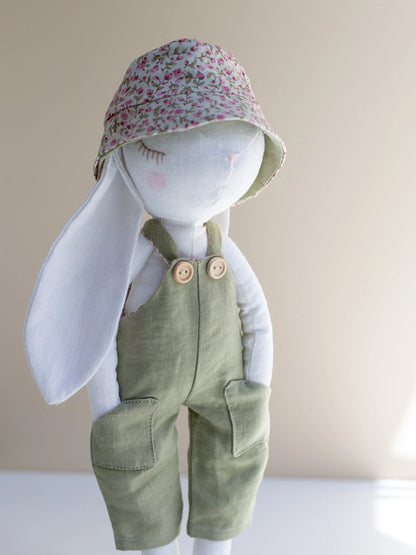 Stuffed bunny toy wearing a floral hat and green overalls on a plain background