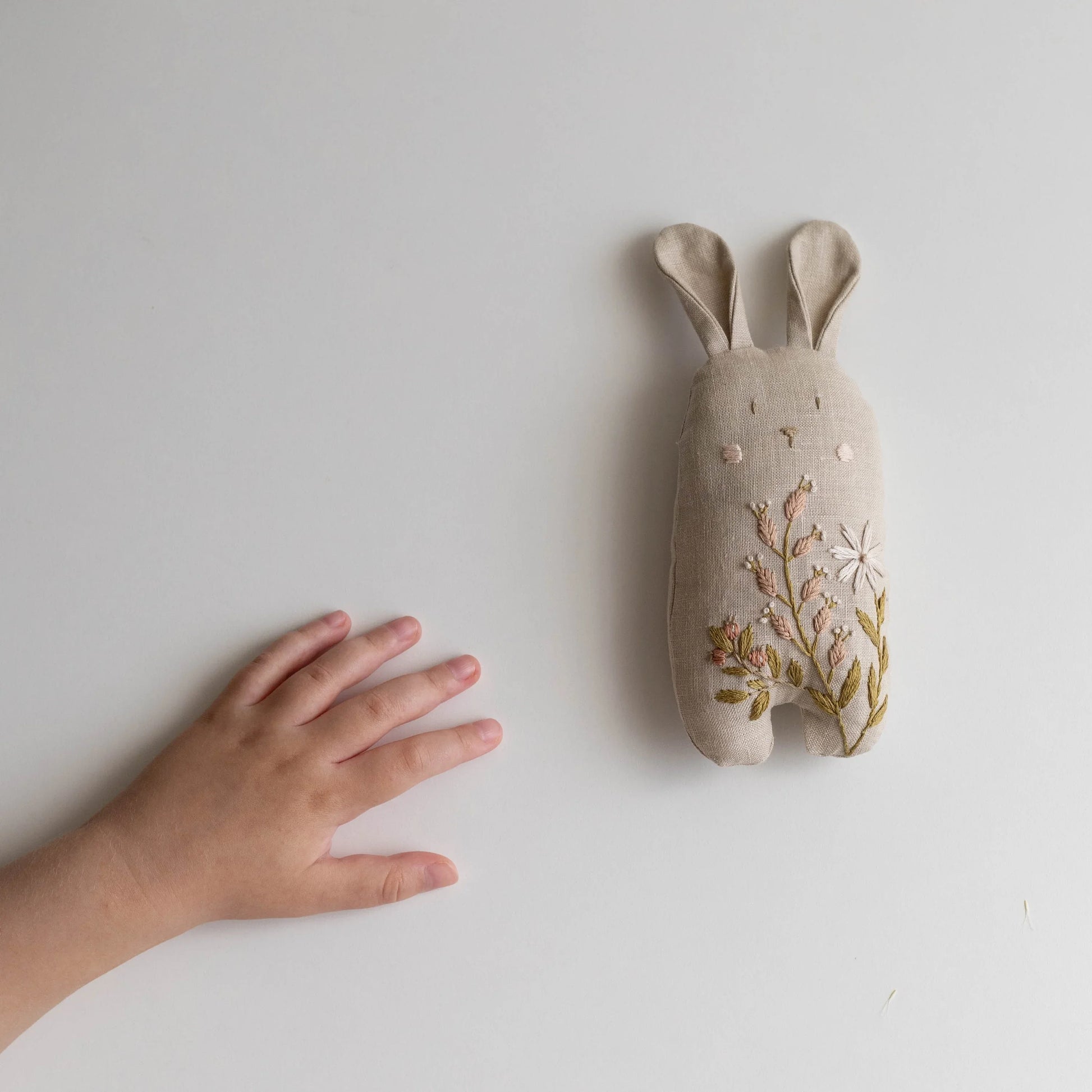Textile rabbit toy with floral embroidery on a white background, next to a child's hand for scale.