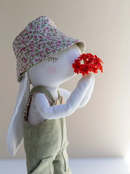Textile art doll wearing a floral hat and green overalls, holding red flowers against a plain background.