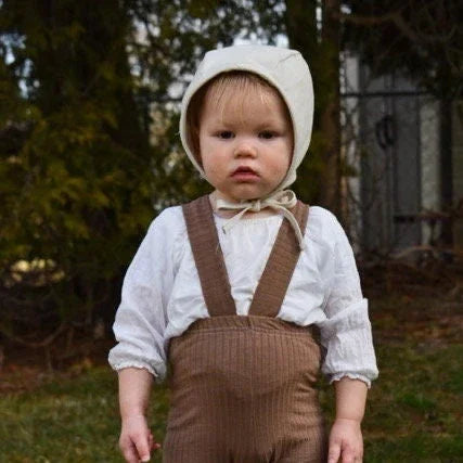 Child wearing a white bonnet and brown overalls standing outdoors.