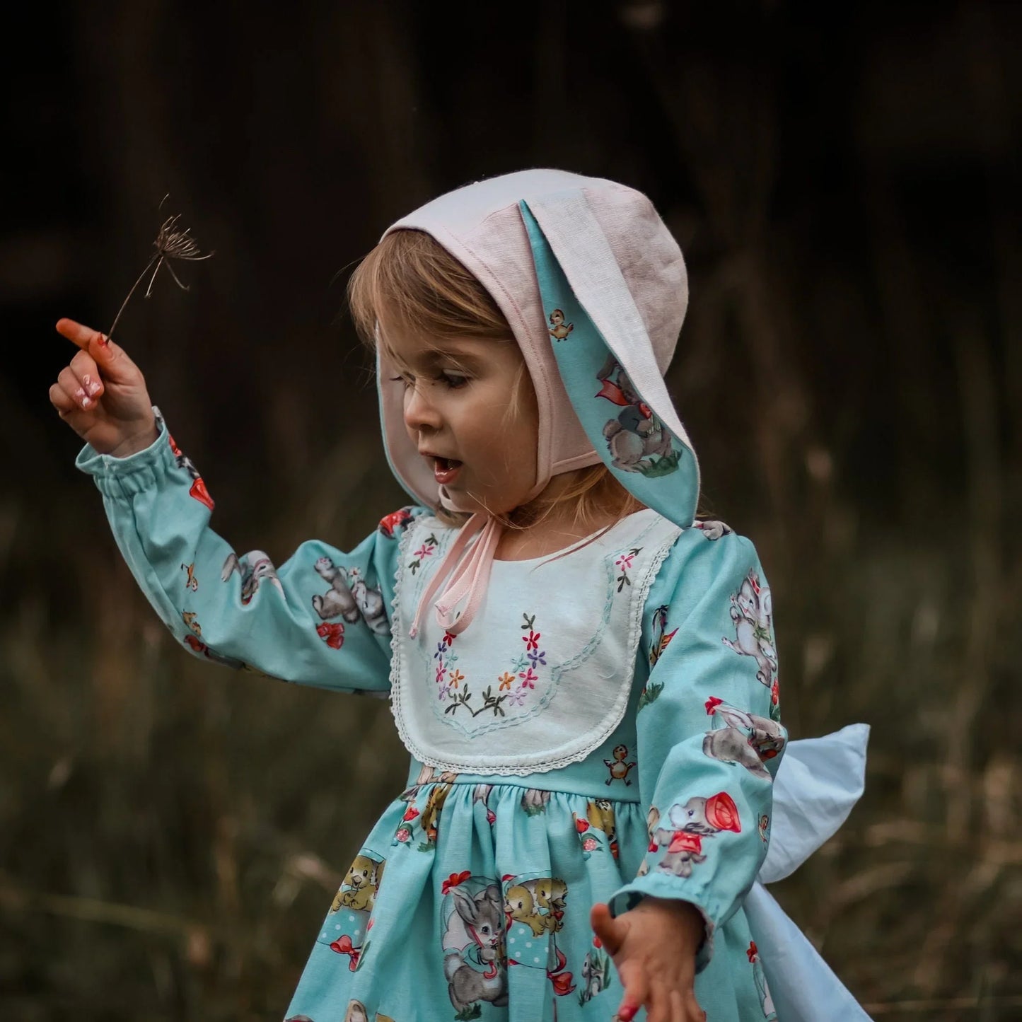 Child in a floral dress and bonnet holding a dandelion seed head outdoors.