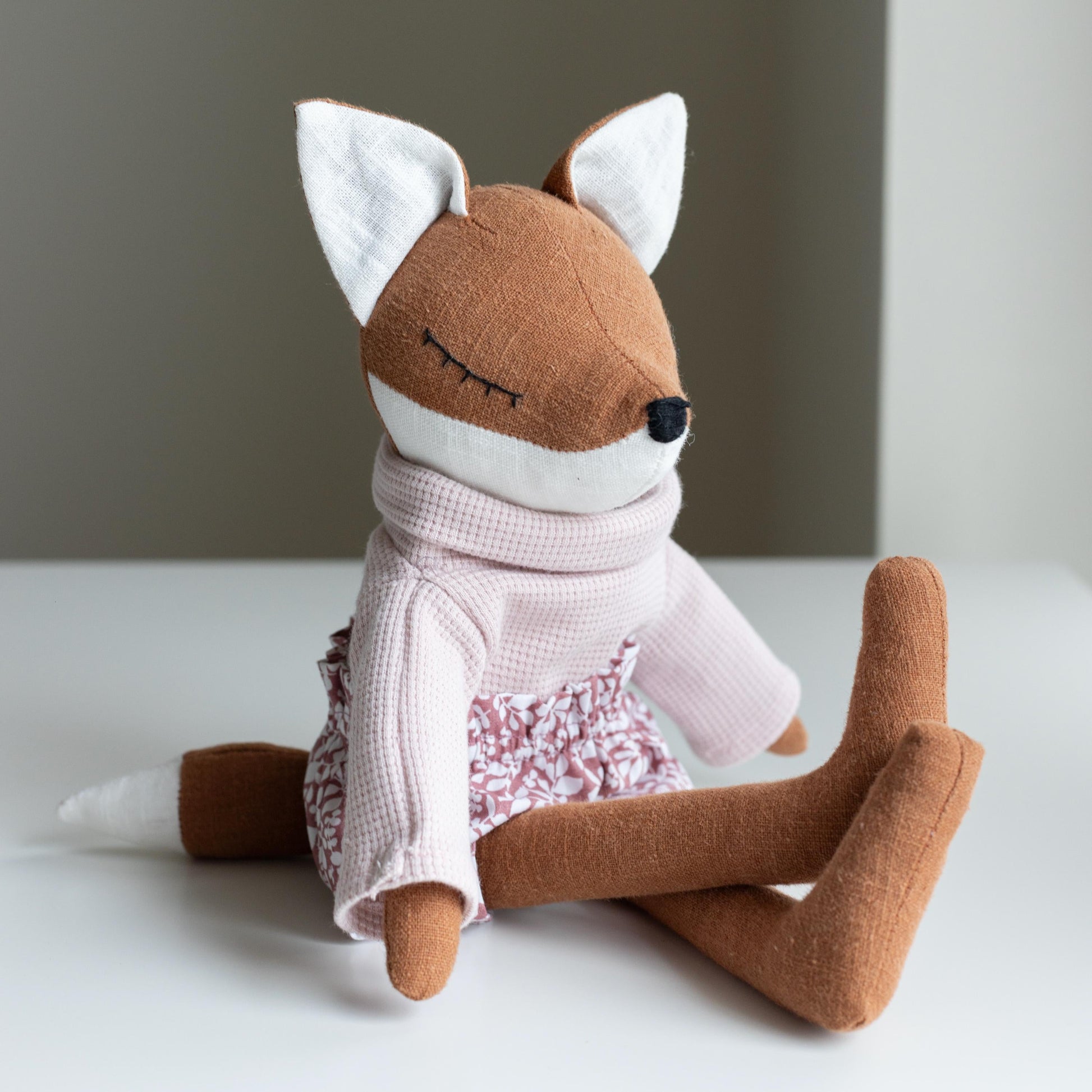 Stuffed fox toy wearing a pink outfit on a white surface with a neutral background