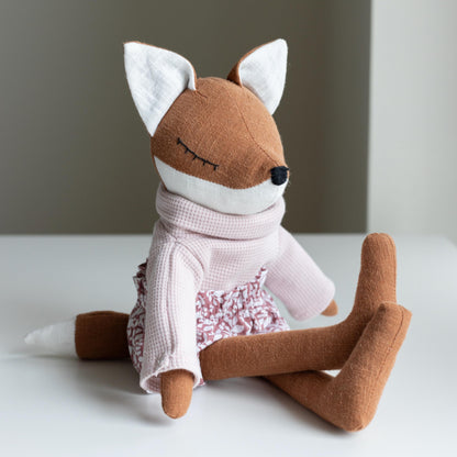 Stuffed fox toy wearing a pink outfit on a white surface with a neutral background