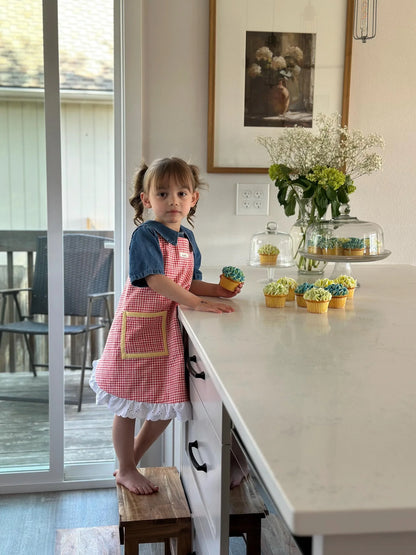 Child in a red apron standing at a kitchen counter with small decorated cakes.