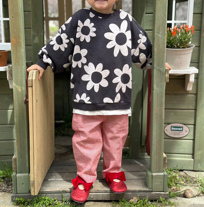 Child wearing a black sweater with white flower patterns, pink pants, and red shoes standing in a wooden playhouse.