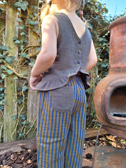 Person wearing a checkered vest and striped pants standing outdoors with greenery and a pot in the background.