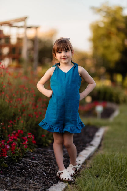 Young girl in a blue dress standing outdoors with flowers and greenery in the background