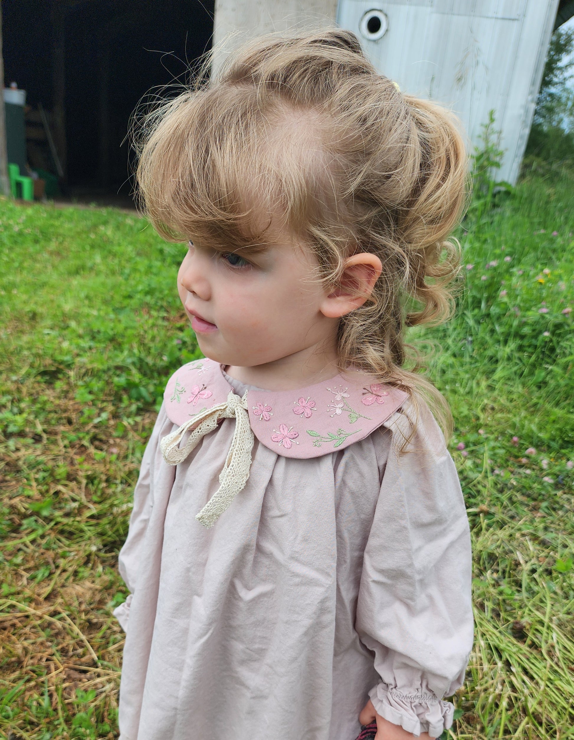 Young girl with styled hair wearing a light-colored dress with a pink collar in an outdoor setting.